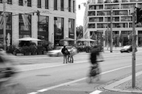 Eine Straßenszene in schwarz-weiß. Menschen gehen und Fahrzeuge fahren vorüber und mittendrin steht oder sitzt jemand, von den anderen ungesehen.