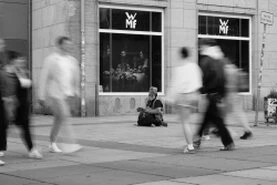 Eine Straßenszene in schwarz-weiß. Menschen gehen und Fahrzeuge fahren vorüber und mittendrin steht oder sitzt jemand, von den anderen ungesehen.