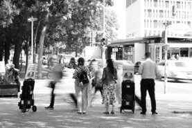 Eine Straßenszene in schwarz-weiß. Menschen gehen und Fahrzeuge fahren vorüber und mittendrin steht oder sitzt jemand, von den anderen ungesehen.