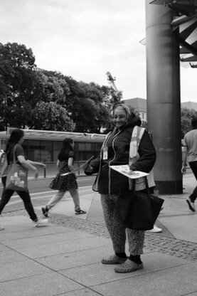 Eine Straßenszene in schwarz-weiß. Menschen gehen und Fahrzeuge fahren vorüber und mittendrin steht oder sitzt jemand, von den anderen ungesehen.
