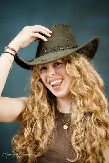 eine lächelnde junge Frau mit langen, blonden, lockigen Haaren und braunem Shirt und einem Cowboyhut auf dem Kopf